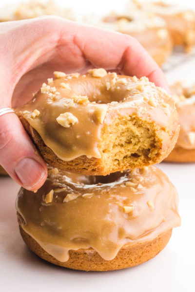 A hand holds a caramel donut hovering over another donut. There are more in the background, too.