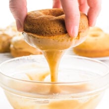 A hand holds a donut over a bowl of caramel glaze. The donut is dripping the glaze after having just being dipped. There are more donuts in the background.