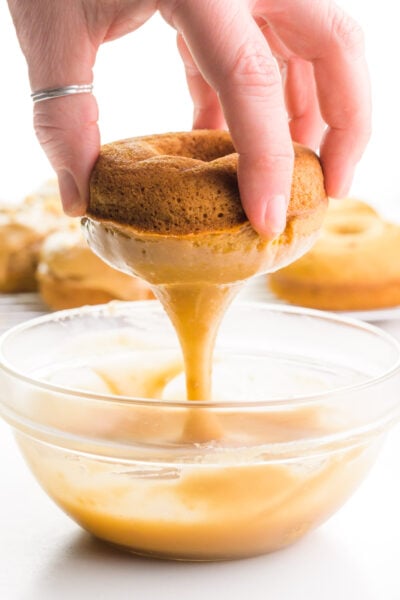 A hand holds a donut over a bowl of caramel glaze. The donut is dripping the glaze after having just being dipped. There are more donuts in the background.