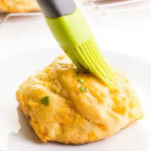 A hand holds a pastry brush, spreading topping over a biscuit. There are more biscuits in the background.