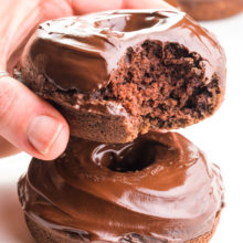 A hand holds a vegan chocolate donut with a bite taken out. It's hovering over another donut and there more in the background.