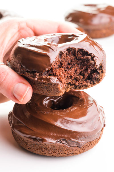 A hand holds a vegan chocolate donut with a bite taken out. It's hovering over another donut and there more in the background.
