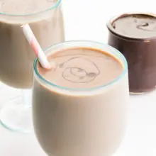 A glass of vegan chocolate milk has chocolate swirls on top and a pink straw on the side. There is another glass and chocolate syrup in the background.