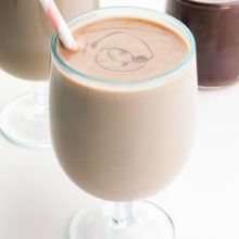 Two glasses of vegan chocolate milk sit one in front of the other, both with pink straws. There is a jar of chocolate syrup in the background.