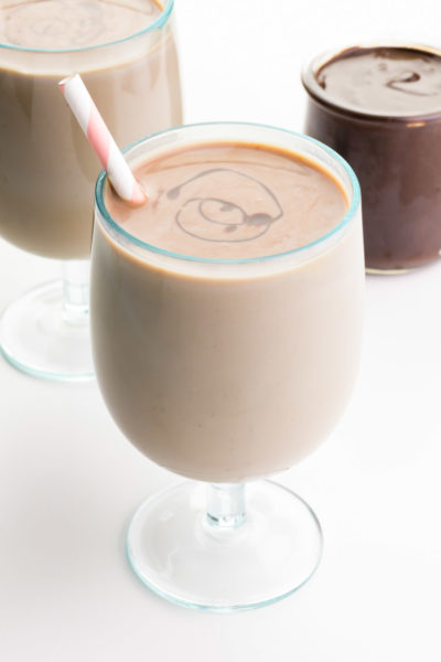 Two glasses of vegan chocolate milk sit one in front of the other, both with pink straws. There is a jar of chocolate syrup in the background.