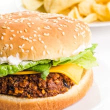 A vegan chorizo burger sits on a plate with a plate of potato chips in the background.