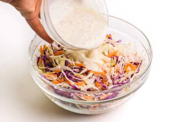 A hand holds a bowl of sauce pouring it over shredded cabbage and carrots.