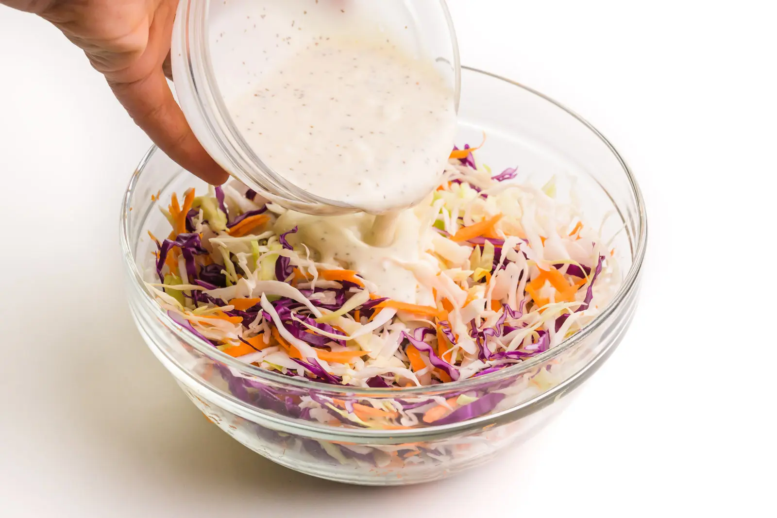 A hand holds a bowl of sauce pouring it over shredded cabbage and carrots.