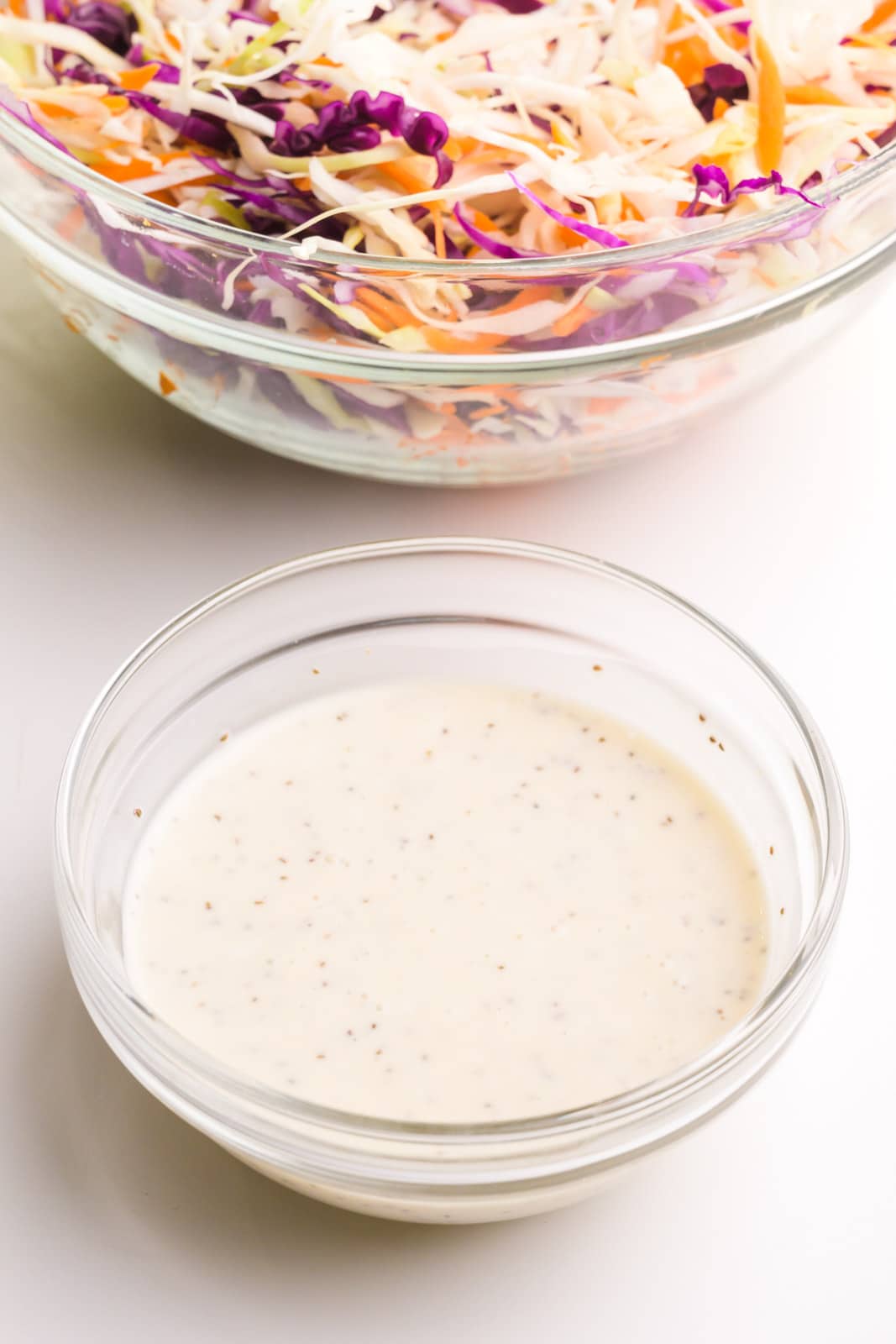 A bowl of creamy coleslaw sauce sits in front of a larger bowl full of shredded cabbage and carrots.