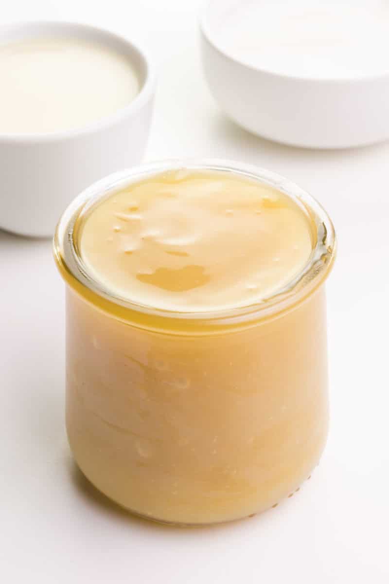 A jar full of vegan sweetened condensed milk sits in front of bowls of sugar and plant-based milk.