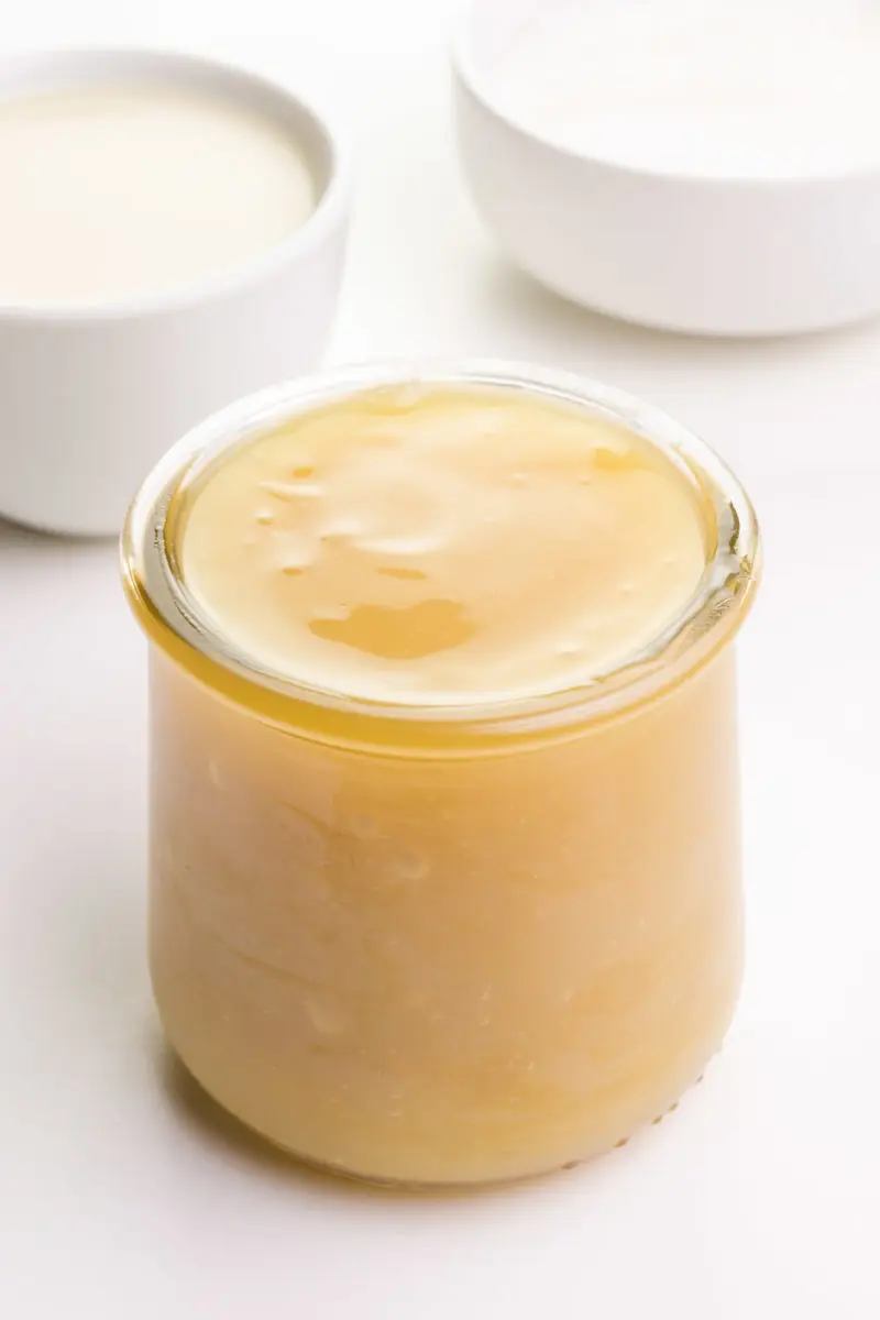A jar full of vegan sweetened condensed milk sits in front of bowls of sugar and plant-based milk.
