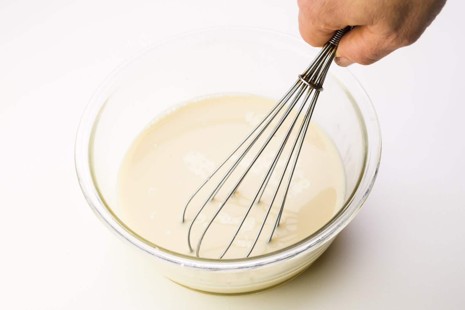 A hand holds a whisk, whisking a milk mixture in a glass pyrex bowl.