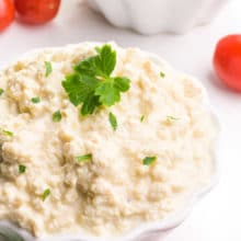 A bowl of vegan cottage cheese has green herbs on top. There's another bowl in the background, along with cherry tomatoes and more fresh herbs.