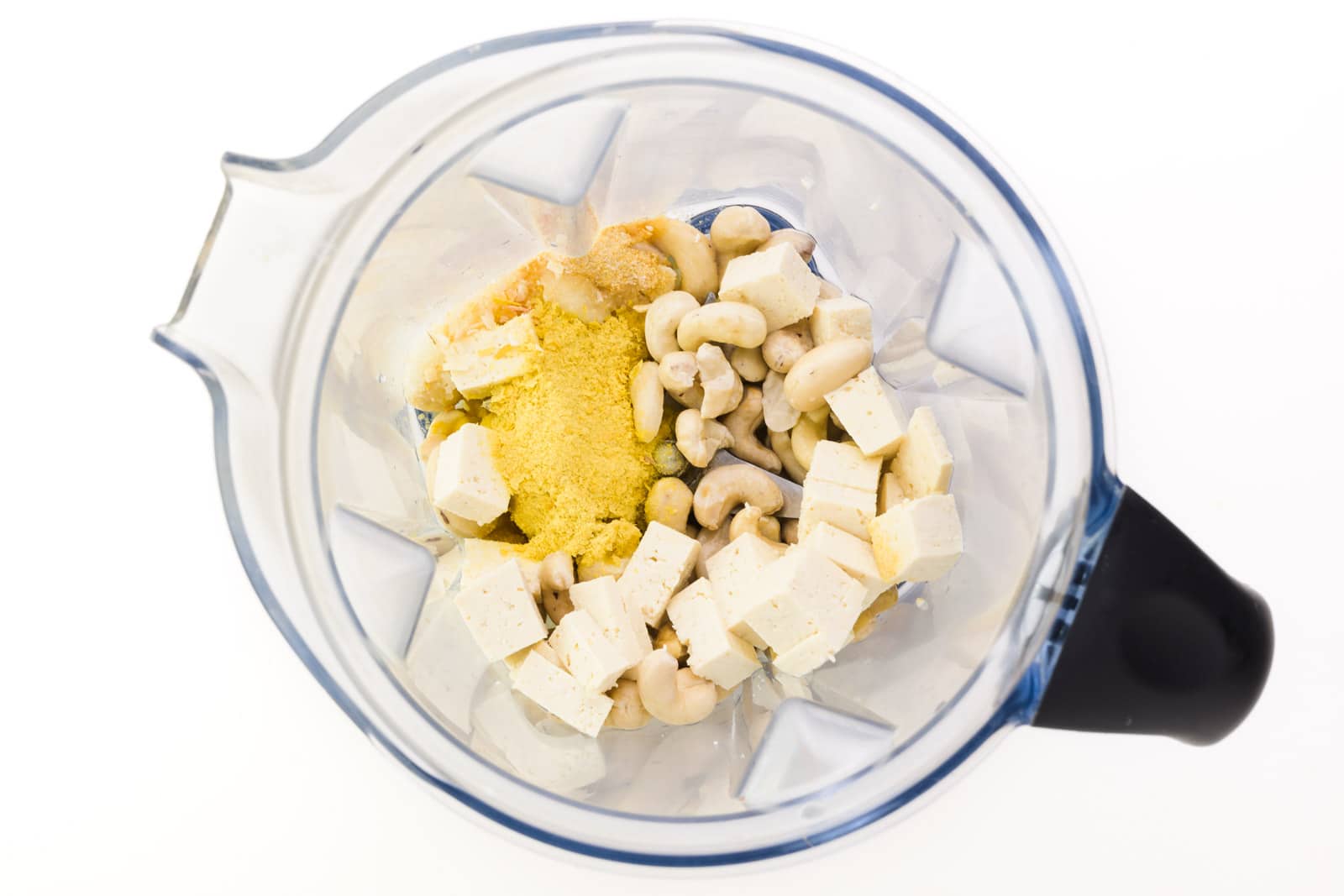 Looking down into a blender jar, there is crumbled tofu, soaked cashews, and more.