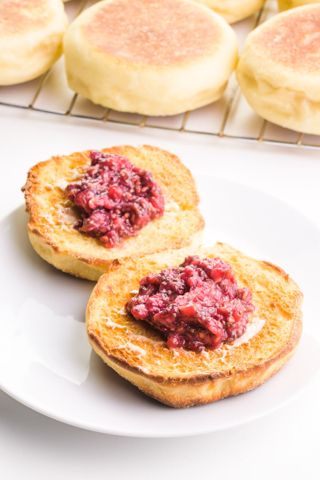 Toasted English muffins have jam on top. There's a wire rack with more in the background.