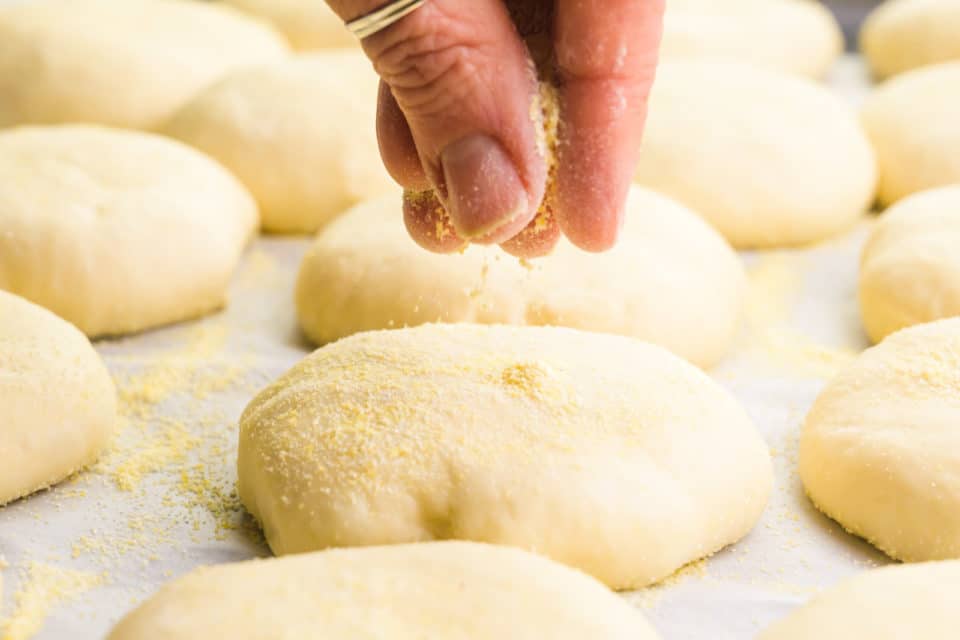 A hand sprinkles cornmeal over flattened dough balls on a baking sheet lined with parchment paper.