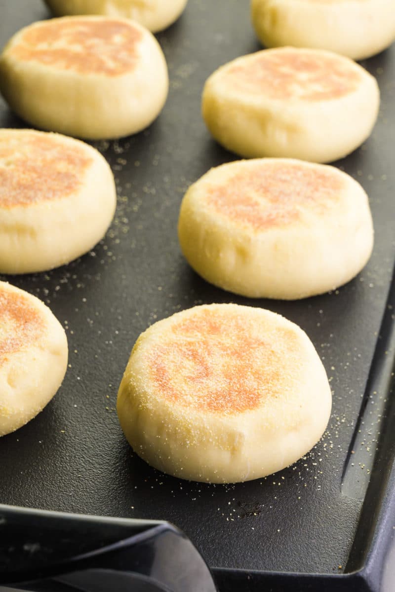 English muffins are cooking on a griddle. The tops are golden-brown.