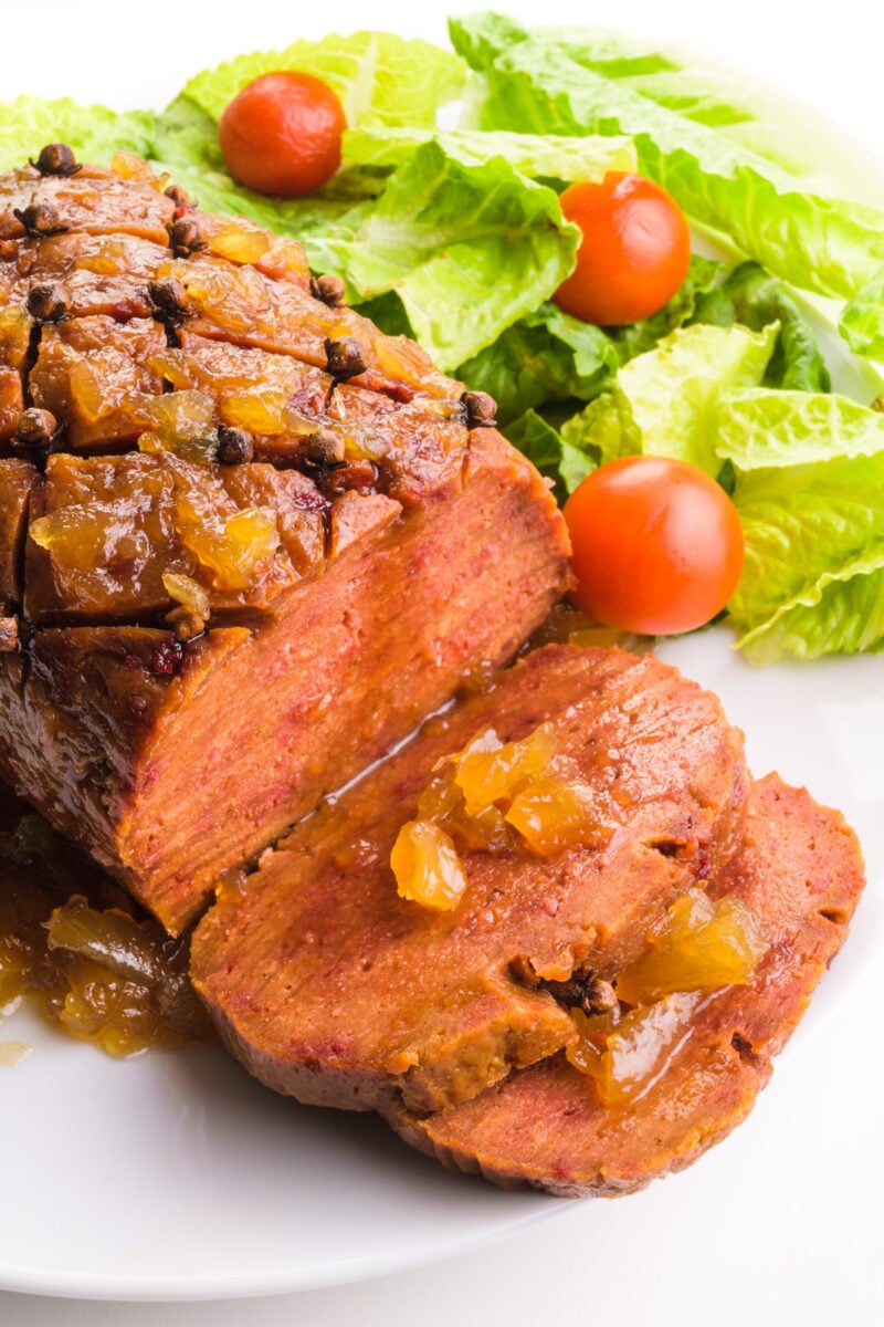 Looking down on a vegan ham with slices cut out. There is pineapple sauce on top of the loaf and on the slices. There is a salad with cherry tomatoes on the back side of the plate.