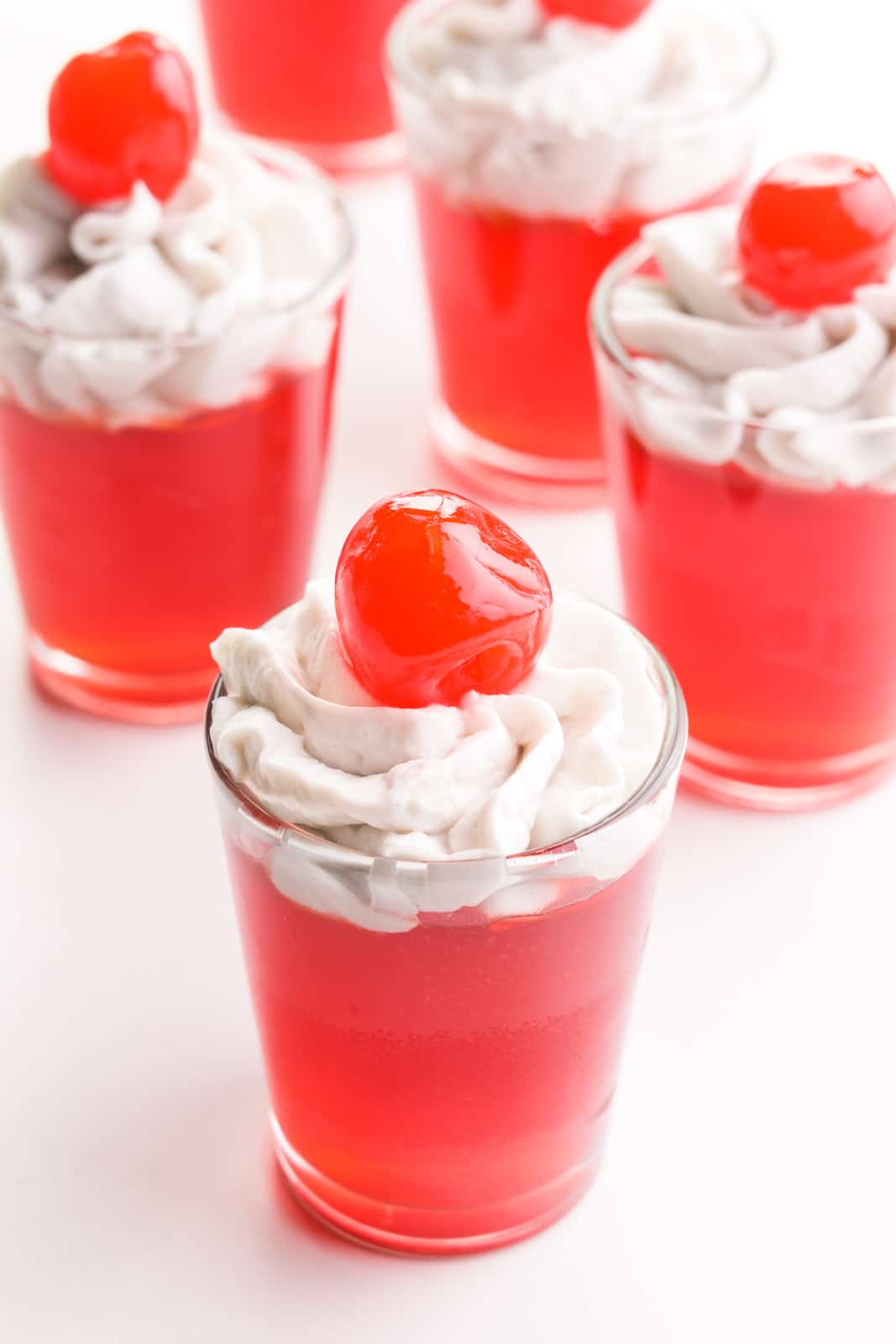 Several jello shots sit next to each other. They each have whipped cream on top and maraschino cherries.