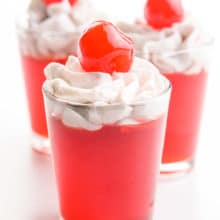 Three shot glasses have cherry vegan jello shots that are topped with whipped cream and maraschino cherries.