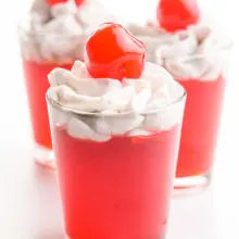 Three shot glasses have cherry vegan jello shots that are topped with whipped cream and maraschino cherries.