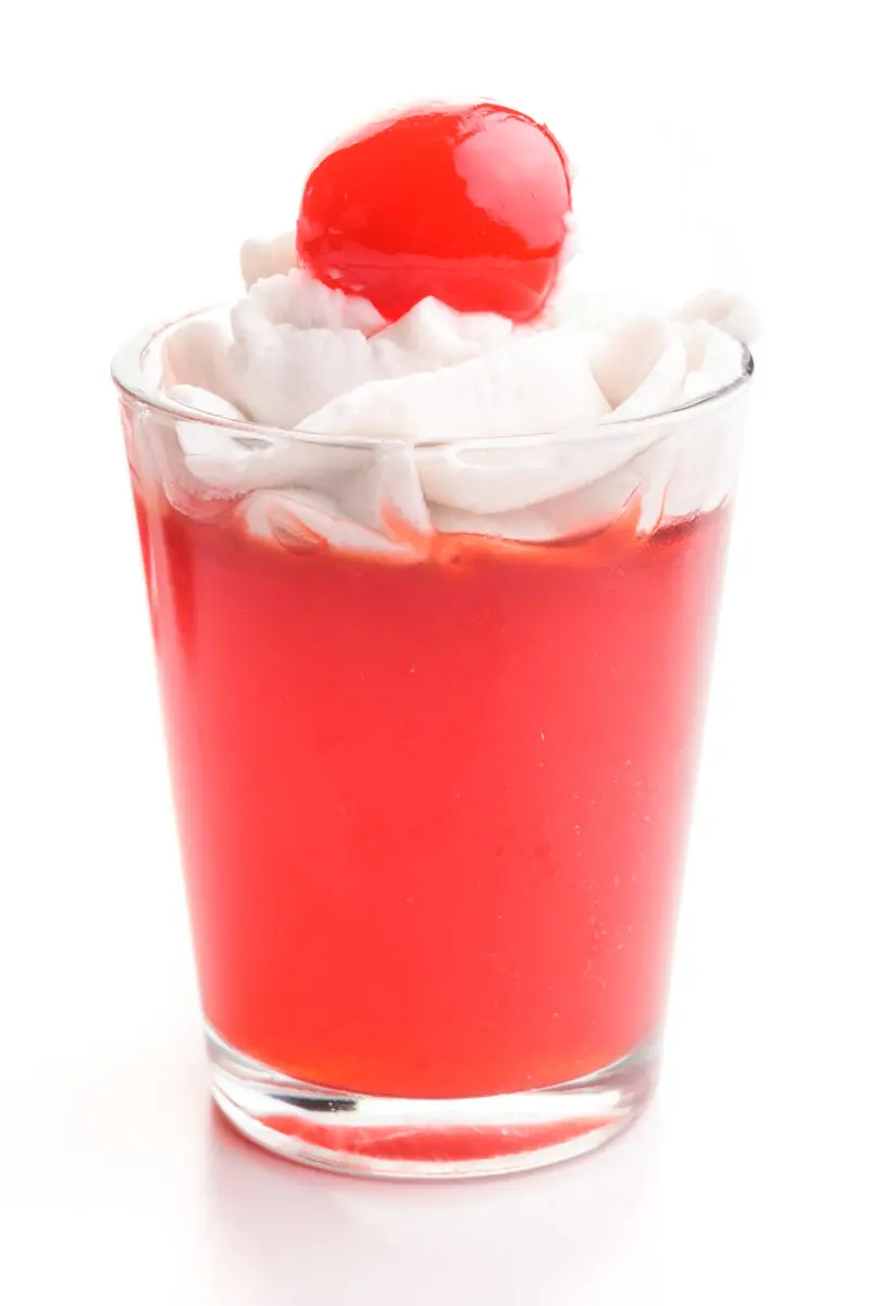 A single cherry jello shot sits on a white table. It has whipped cream and a cherry on top.
