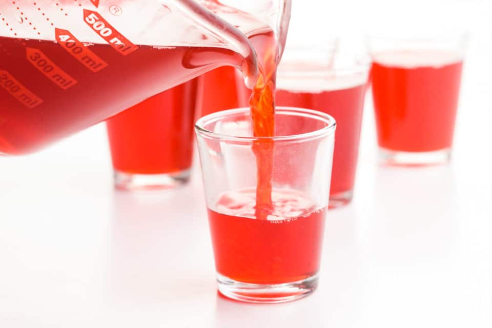 Red liquid is being poured from a glass pyrex measuring cup into jello shots.
