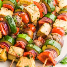Cooked tofu skewers are on a plate. One skewer is placed diagonally across the others. There are pieces of fresh parsley and cherry tomatoes around the plate.