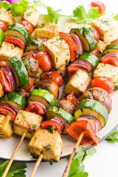 Cooked tofu skewers are on a plate. One skewer is placed diagonally across the others. There are pieces of fresh parsley and cherry tomatoes around the plate.