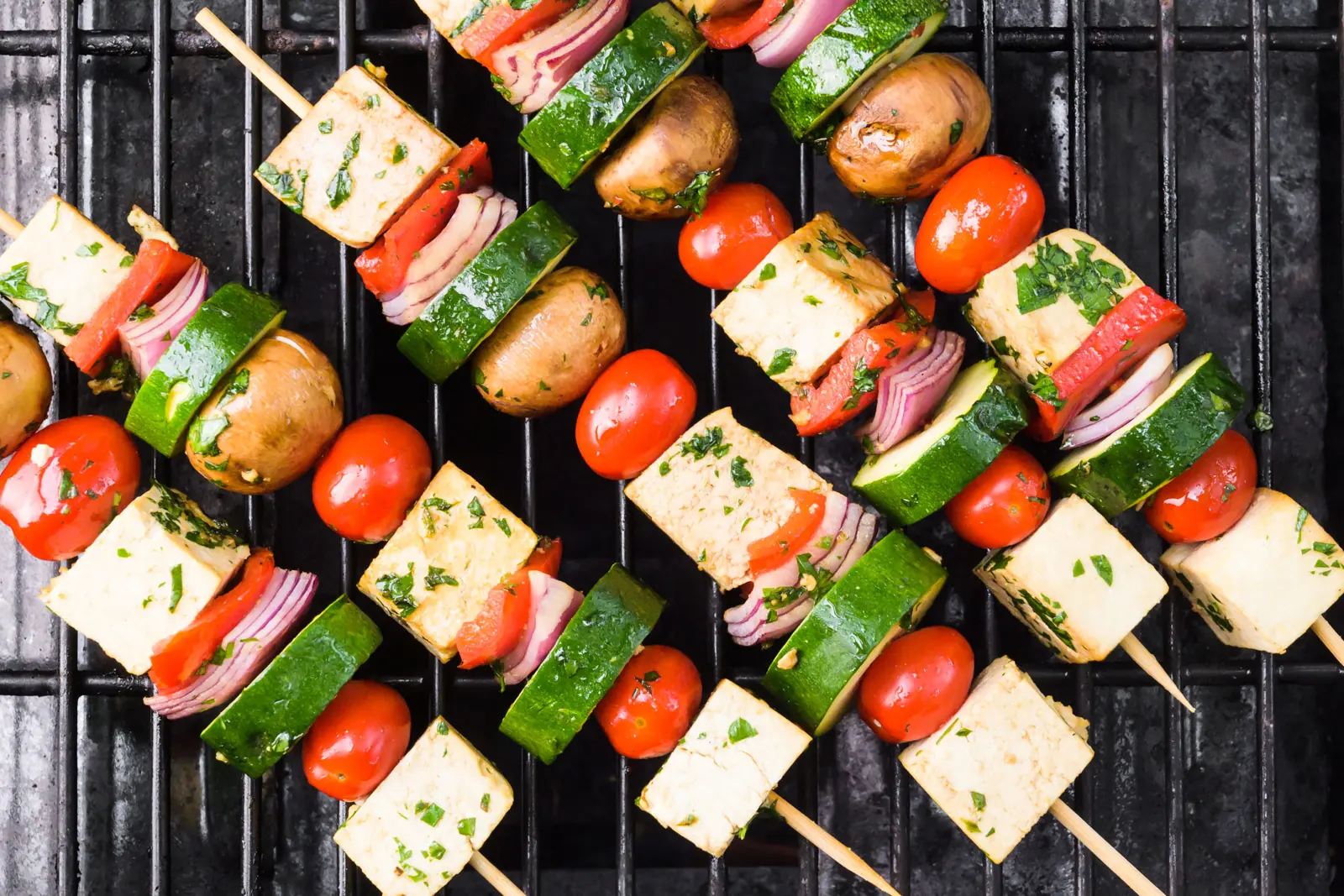 Looking down on vegan skewers on the grill. There are tofu cubes, sliced zucchini, cherry tomatoes, and more.