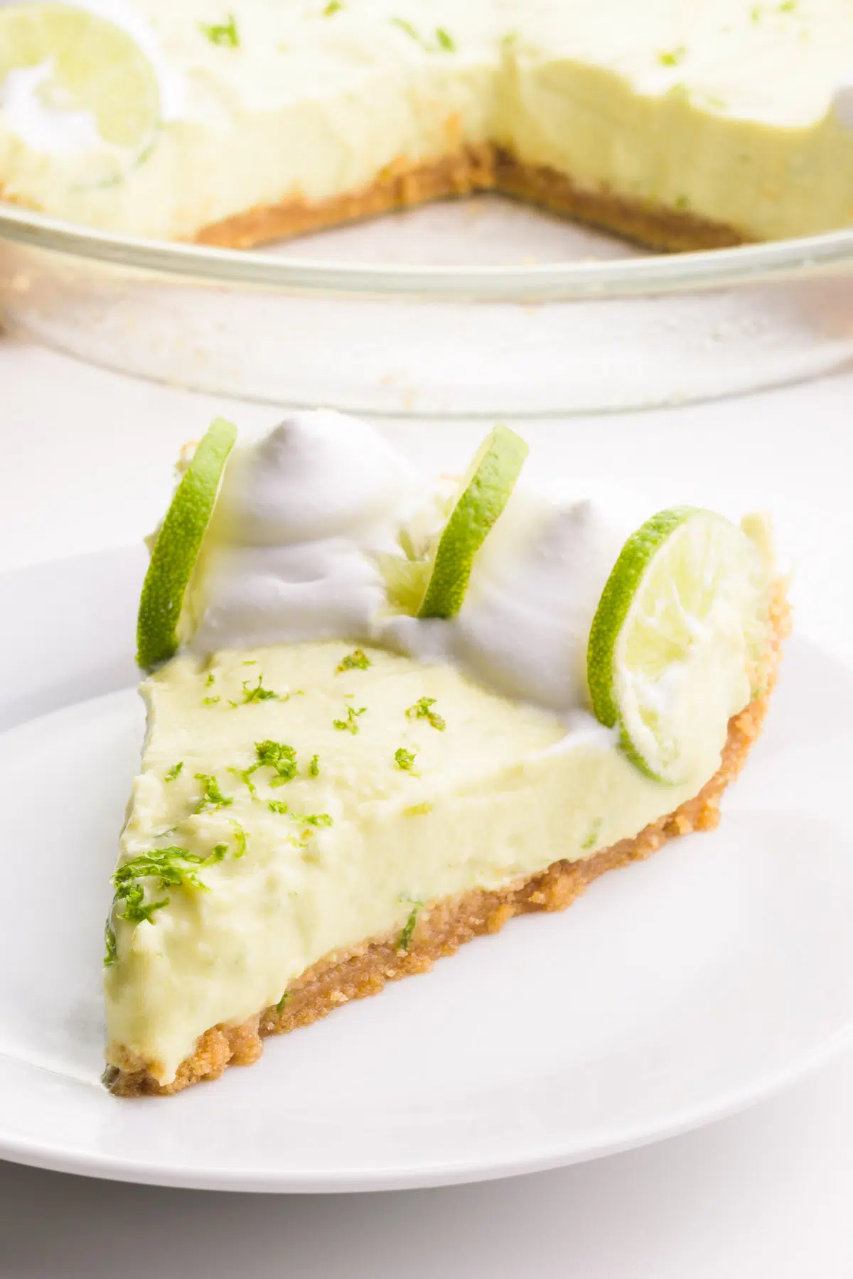 A slice of vegan key lime pie sits on a plate. It has whipped cream and lime slices on the pie. The rest of the pie is in the background.