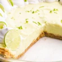 A key lime pie has several slices cut out, revealing the crust and filling. There's whipped cream and lime slices on top of the pie.