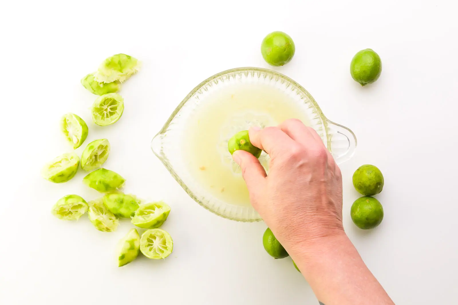 A hand holds a key lime on a glass zester. There are fresh key limes around it along with several key limes that have already been juiced.