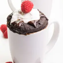 Two white coffee mugs hold chocolate vegan mug cakes with chocolate syrup, whipped cream, and raspberries on top. There are fresh raspberries on the table next to the mugs.