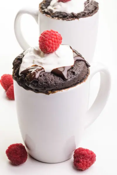 Two white coffee mugs hold chocolate vegan mug cakes with chocolate syrup, whipped cream, and raspberries on top. There are fresh raspberries on the table next to the mugs.
