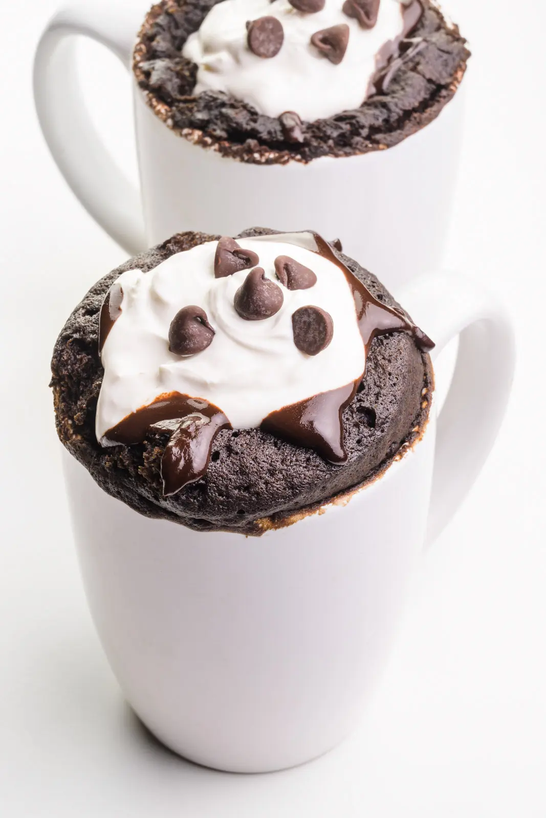 Two chocolate mug cakes have chocolate syrup, whipped cream, and chocolate chips on top.