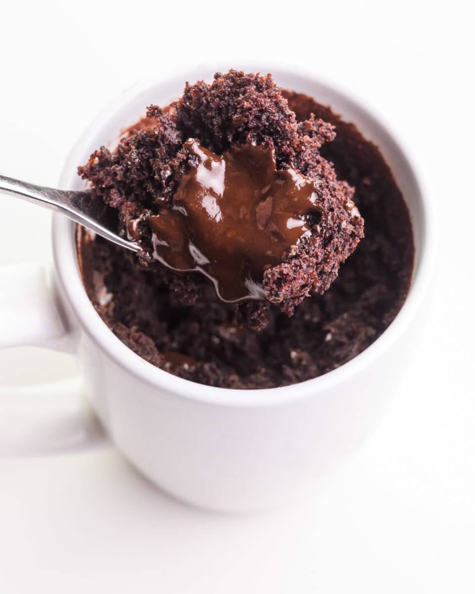 A spoon holds a bite of chocolate mug cake with chocolate syrup on top. it's hovering over the mug with the rest of the cake.