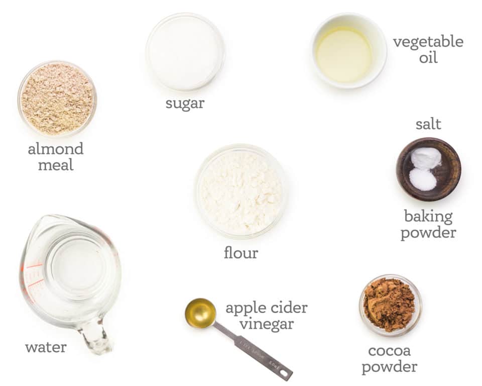 Ingredients are laid out on a white table top. The labels next to each ingredient reads, vegetable oil, salt, baking powder, cocoa powder, apple cider vinegar, sugar, water, and flour.