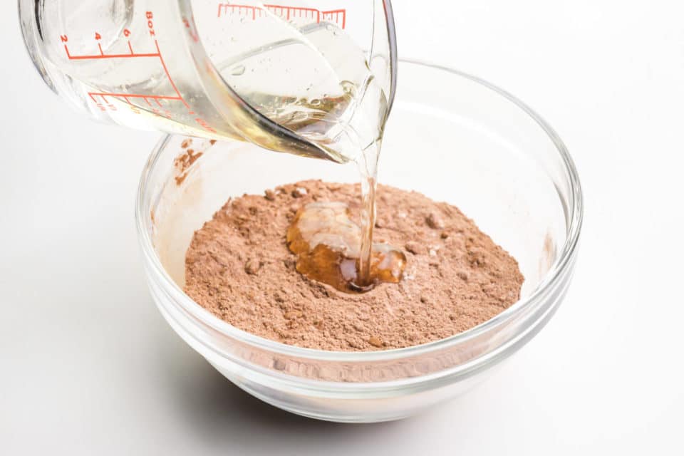 A water mixture is being poured from a glass pyrex measuring cup into a bowl with a cocoa powder mixture.