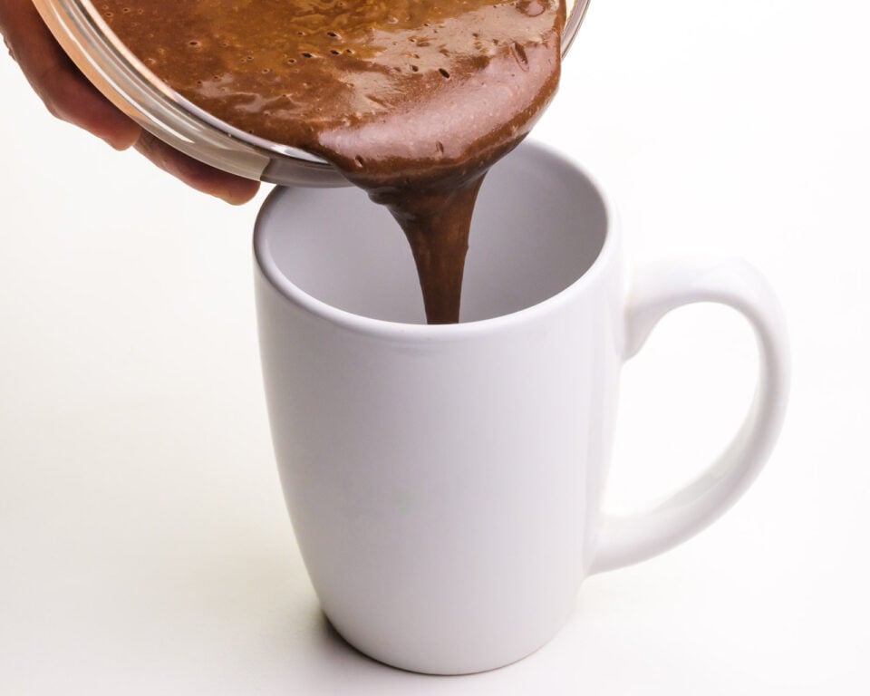 A hand holds a bowl of chocolate cake batter pouring it into a white coffee mug.
