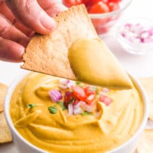 A hand holds a tortilla chip with lots of cheese sauce on it, hovering over the bowl with more of the sauce. There are more tortilla chips and veggies in the background.