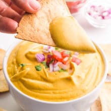 A hand holds a tortilla chip with lots of vegan nacho cheese sauce on top. The bowl of the sauce is below it and there are other ingredients like chopped tomatoes and red onions on top.