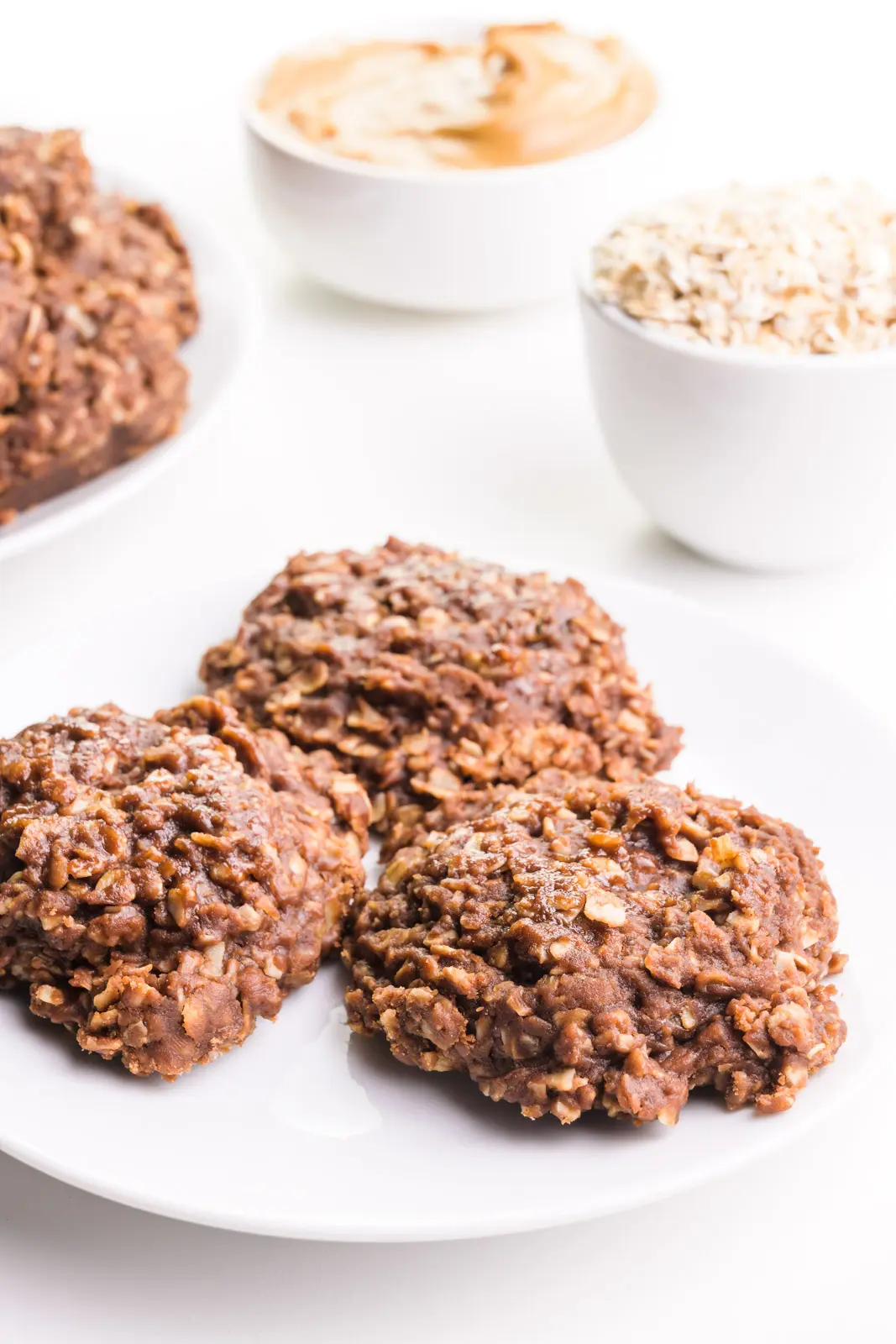 Three chocolate no-bake cookies are on a plate. There's a bowl of oats, a bowl of peanut butter, and a plate with more cookies in the background.