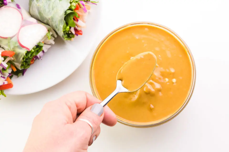 A hand holds a spoon hovering over a bowl of peanut sauce. A plate sits nearby holding veggie spring rolls.