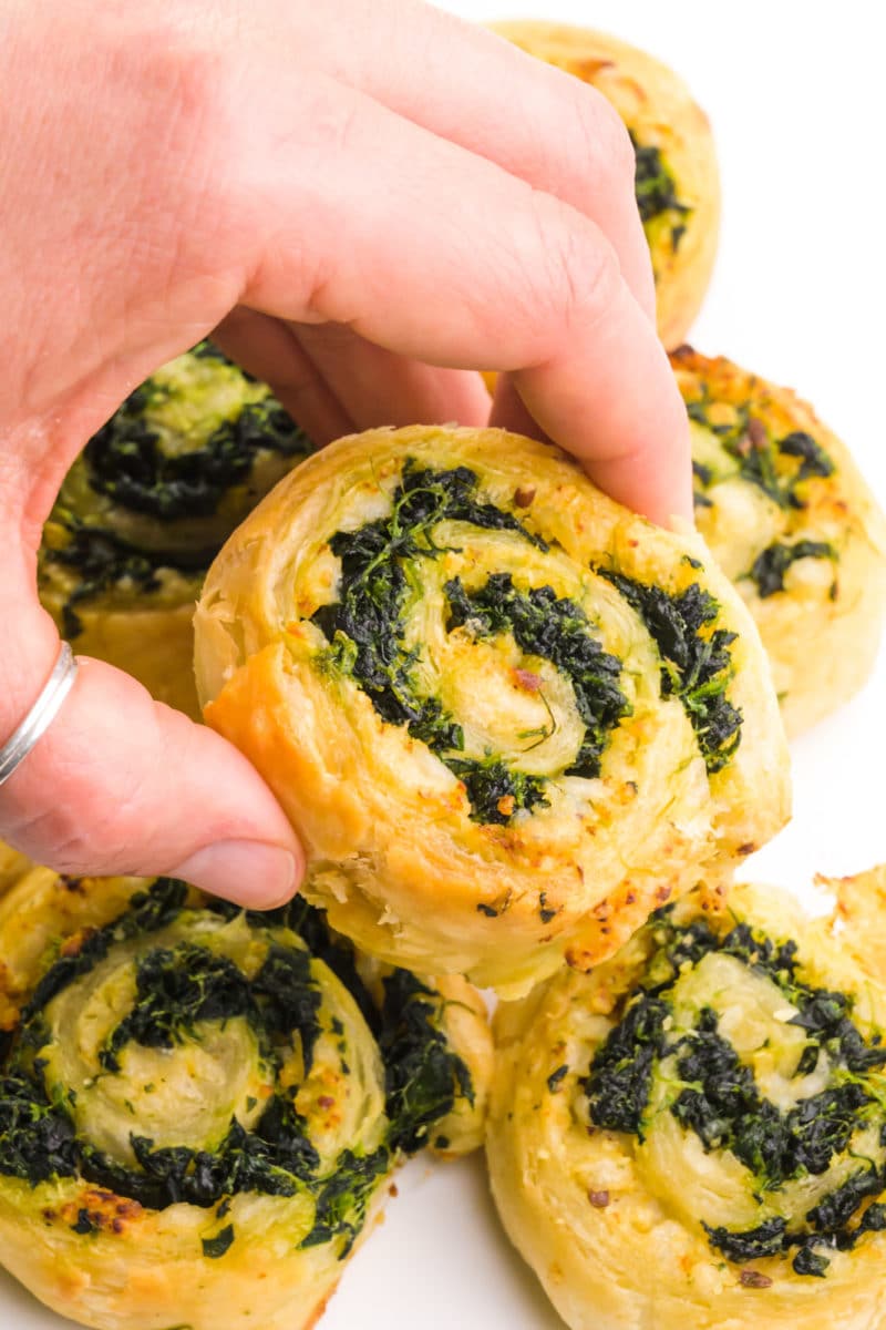 A hand holds a spinach pinwheel, hovering over more of them.