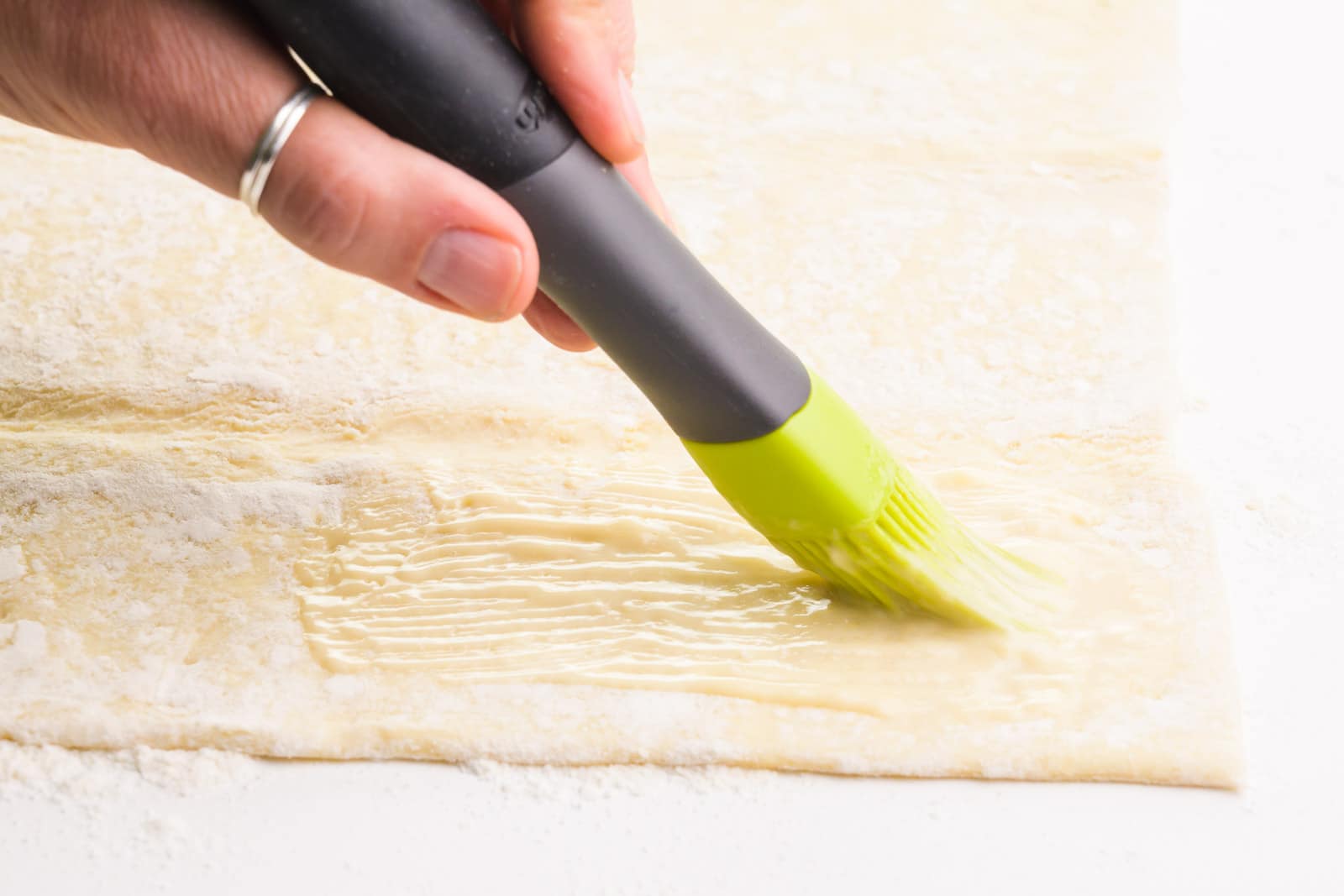 A. hand holds a pastry brush, spreading melted butter over a pastry sheet.