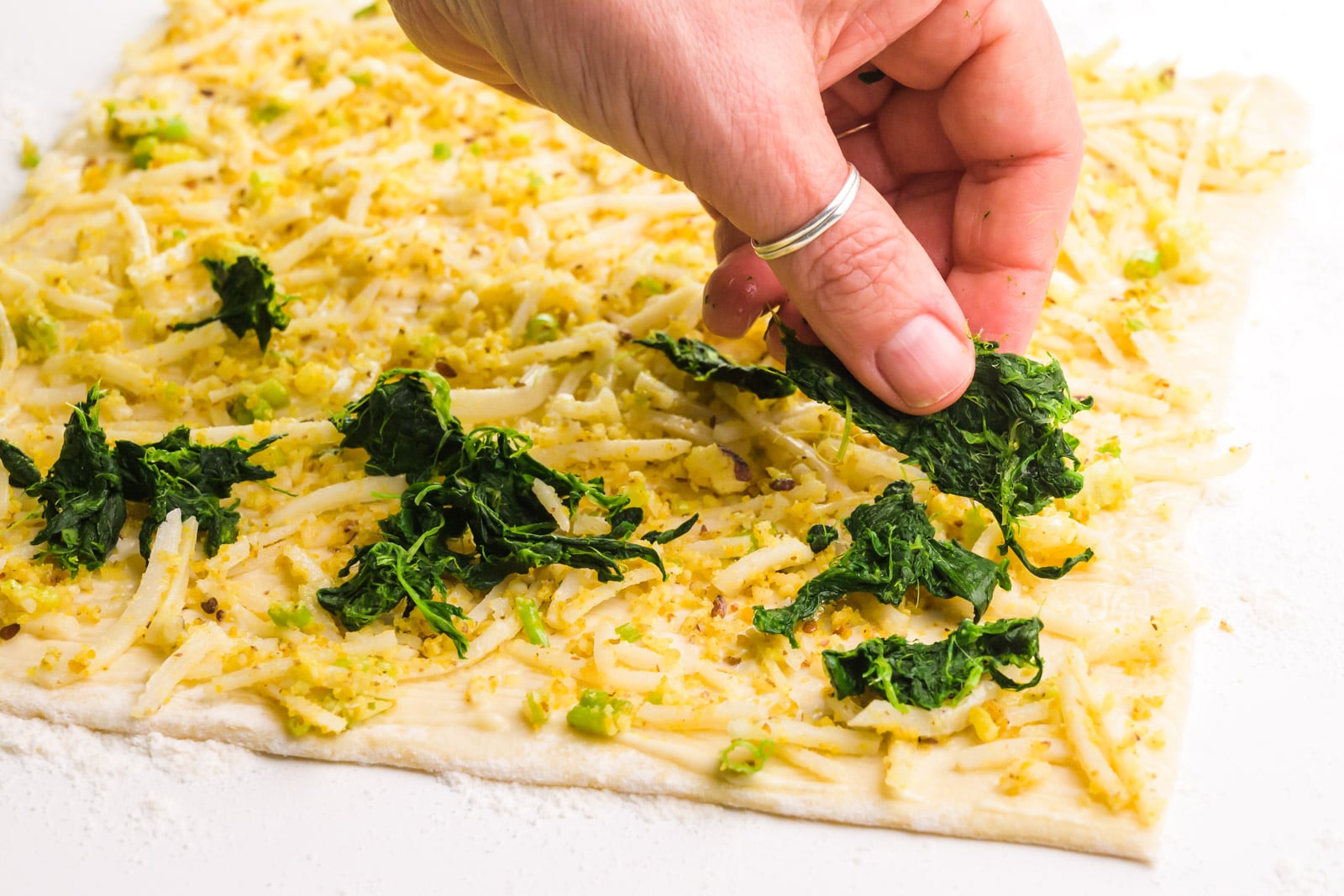 A hand distributes spinach pieces over a pastry sheet lined with cheese and butter.