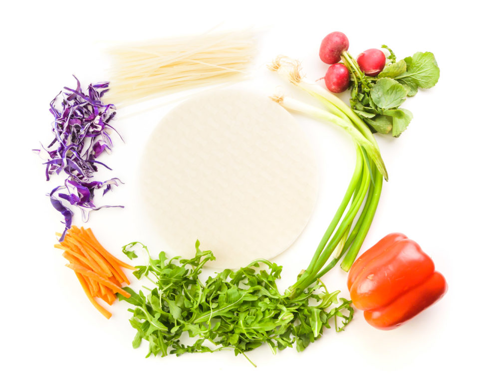 Looking down on ingredients on a white counter, including rice papers, radishes, green onions, a red bell pepper, greens, carrot sticks, purple cabbage, and uncooked rice noodles.