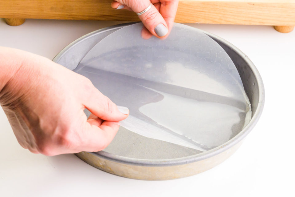 Two hands hold a rice paper and are dipping it in a cake pan full of water. There's a wooden cutting board in the background.