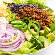 A bowl holds a salad with lots of toppings, such as chopped guacamole, taco veggie crumbles, black beans, vegan cheddar cheese, and more. There is a head of lettuce and tomatoes in the background.